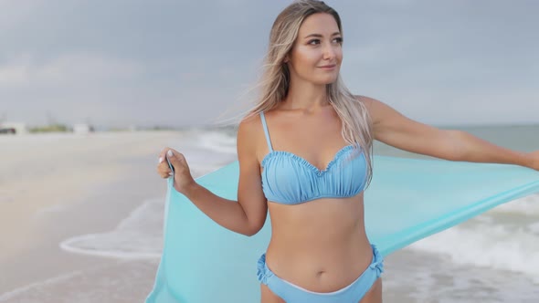 A Girl with Blond Hair in a Bluish Swimsuit and a Bright Shawl Walks Along the Beach