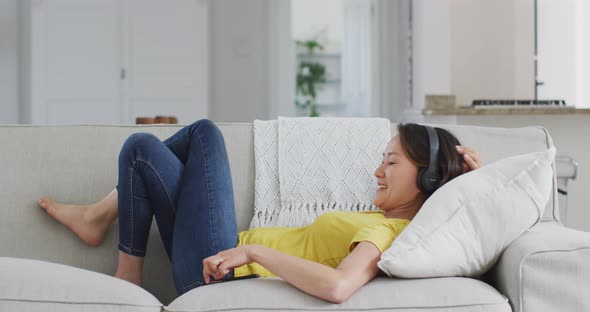 Happy asian woman wearing headphones lying on sofa, resting with smartphone at home