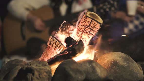 Blurred group of people having fun sitting near bonfire outdoors at night playing guitar 