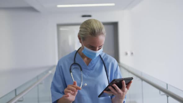 Caucasian female doctor wearing face mask using tablet, looking to camera in hospital corridor