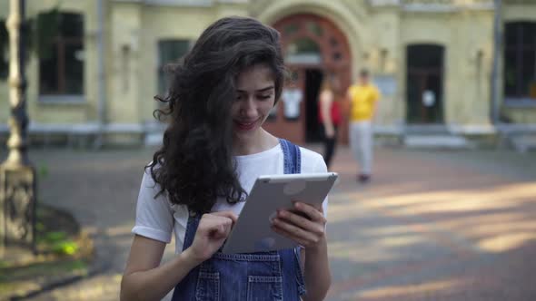 Cheerful Young Woman Messaging Online on Tablet with Blurred Couple of Groupmates at University