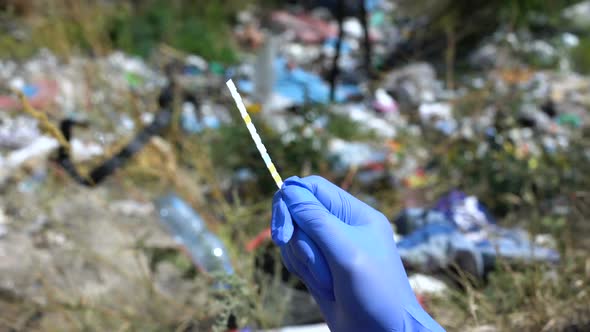 Hand in Glove Holding Ph Test Strip Against Landfill Background, Pollution Level