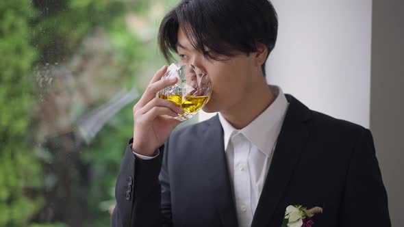 Portrait of Thoughtful Groom in Wedding Suit with Boutonniere Drinking Whiskey From Glass Looking