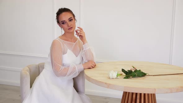 A Slender Beauty in a Wedding Dress Poses Sitting in an Armchair at the Table