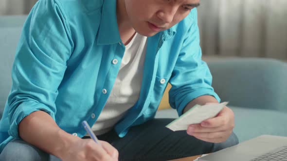 Asian Man With A Laptop Holding And Looking At The Bill While Recording The Expenses In Notebook