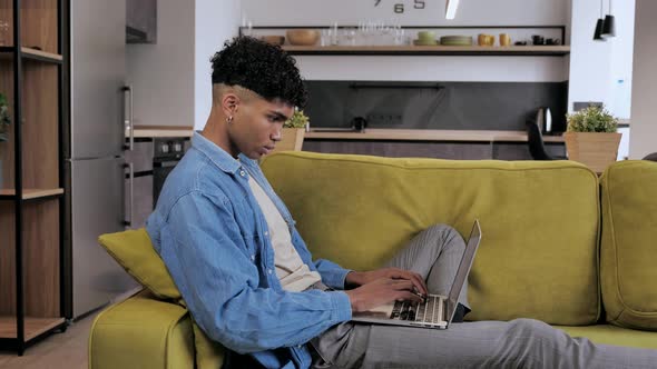 Relaxed Serious Mixed Race Male Teen Freelancer Using Laptop Device Leaning on Sofa at Home Office