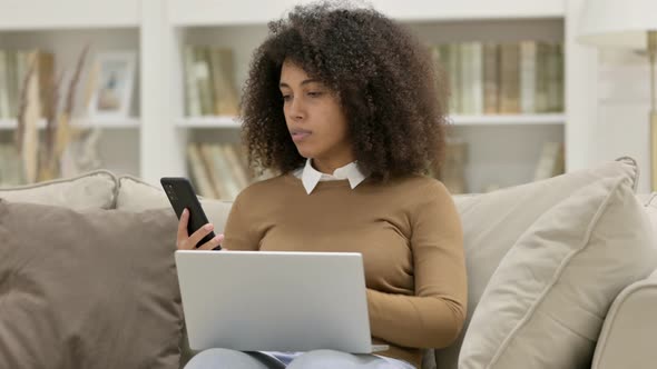 Young African Woman Working on Smartphone and Laptop on Sofa