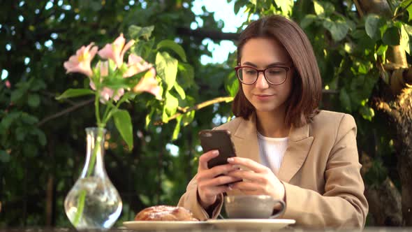 A Young Business Woman Is Sitting in a Cafe and Texting on the Phone. Girl with Coffee and a Bun on