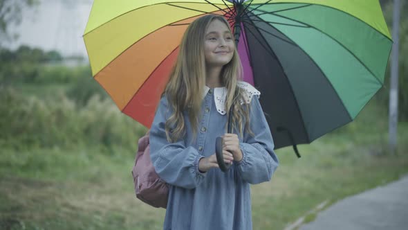 Charming Cheerful Schoolgirl Standing Outdoors with Colorful Umbrella and Smiling