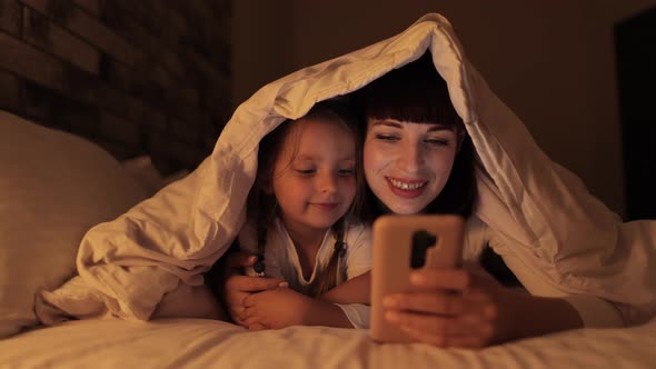 Mom Lying on Bed Under Blanket with Her Daughter Using Mobile Application Spending Evening Time