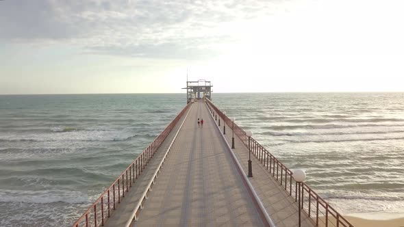 Aerial view of teenagers boy and girl walk by hand on pier in sea foaming waves love on beach summer