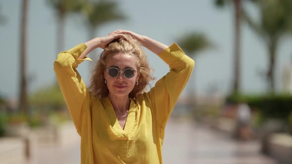 Portrait of a Blonde in Sunglasses and a Yellow Dress Against a Background of Blurred Palm Trees on