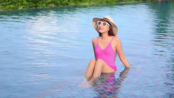 Young attractive Asian woman in pink swimsuit and white sunglasses sitting on edge of pool enjoying