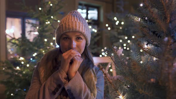 Portrait of a Happy Woman Against the Backdrop of Christmas Decorations. She Rubs Her Hands, Keeping