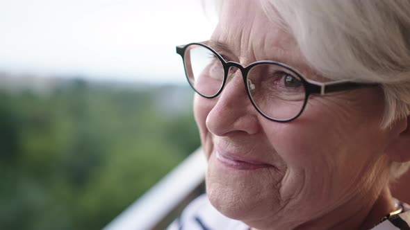 Senior Gray Haired Woman with Glasses Standing Alone on the Balcony with Smile but Sad Eyes