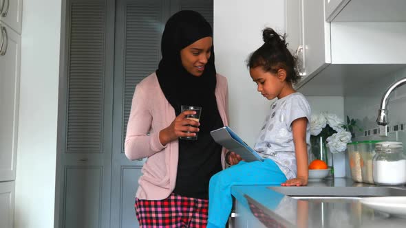 Young mother with a hijab drinking a glass of water near her daughter in the kitchen 4k