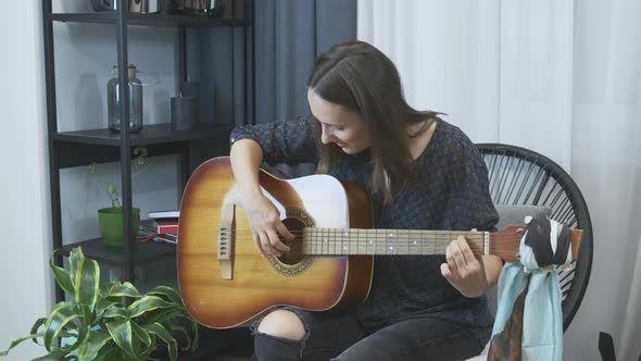 Young attractive girl playing acoustic guitar in cozy chair at modern home studio.