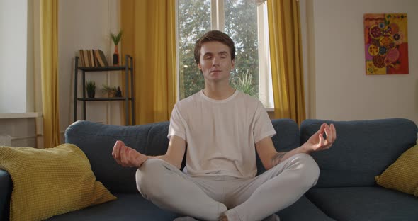 Young Man Sitting at Sofa Practicing Yoga Relaxation After Busy Workweek Slow Motion. Front View of