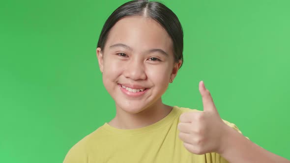 Happy Young Asian Kid Girl Showing Thumbs Up Gesture While Standing In The Green Screen Studio