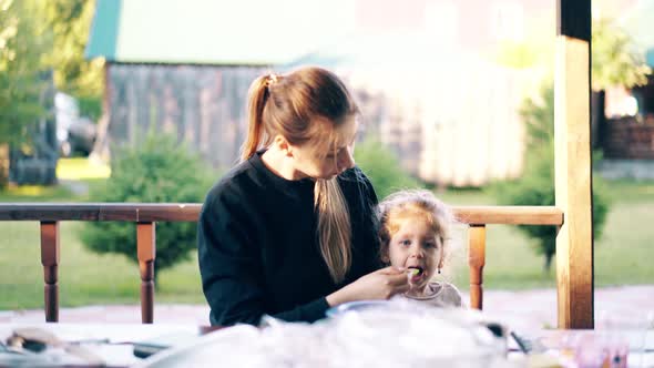 Young Pretty Mother Is Feeding Her Daughter with Porridge, They Are Sitting in a Summer House