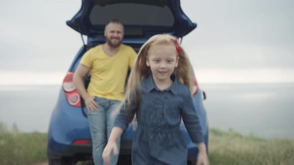 Cute Cheerful Little Girl Running Along Summer Meadow on River Bank with Blurred Father in Car Trunk