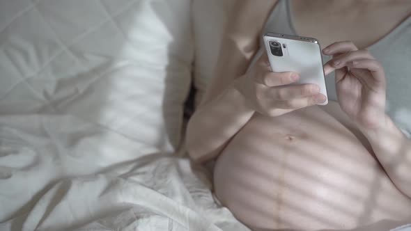 A Pregnant Woman Uses the Phone While Relaxing in a Home Bed