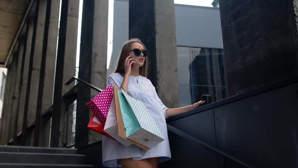 Girl Staying Near Shopping Mall with Shopping Bags and Talking on Mobile Phone About Purchases