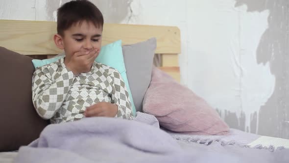 Sick Little Boy Coughing While Sitting in Bed and Covering Himself with a Blanket