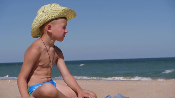 Boy Sits on the Golden Sand. The Child Wears a Straw Hat with a Large Brim