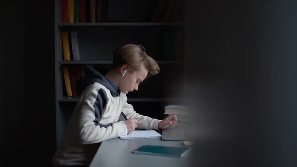 Thoughtful Pupil Boy Learning Online Using Laptop Making Notes in Copybook Sitting at Desk at Home