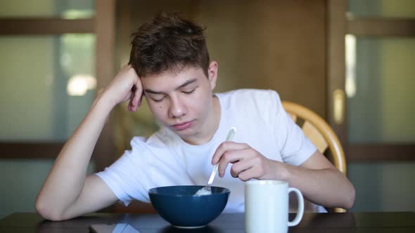 A sleepy teenager lazily eats porridge early in the morning