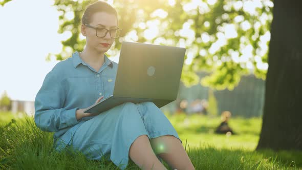 Busy Attractive Woman Working at the Laptop As Sitting on Grass in City Park at Sunset