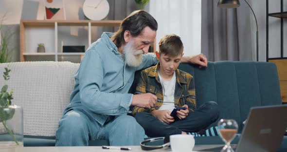 Boy Sitting on the Sofa in Cozy Room Together with His Bearded Grandfather and Use Smartphone