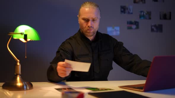 Confident Serious Cop Sitting at Table with Laptop Comparing Photos Thinking