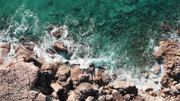 Aerial top view of sea coast line with ocean waves hitting rocky seashore creating white foam 