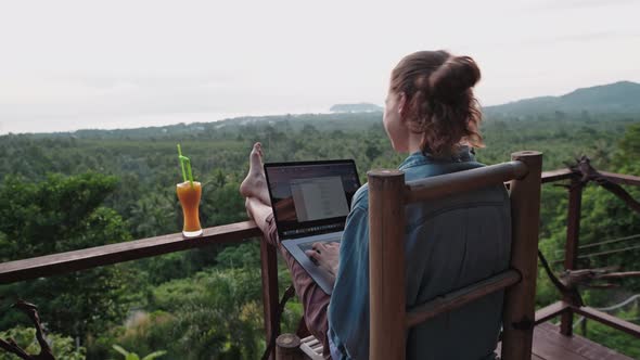 Freelancer woman sits with a laptop on a high bar chair.