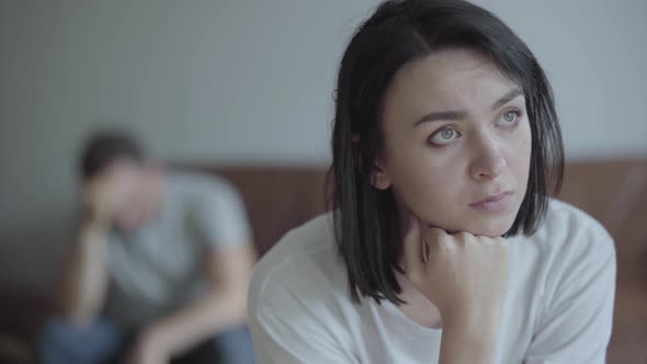 Close-up Portrait of Sad Broken Woman on the Foreground and Her Husband Sitting on the Sofa