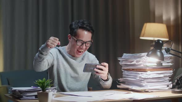 Happy Asian Man Celebrating Winning Game On Smartphone After Working With Documents At Home