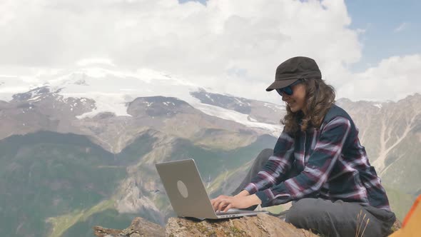 Lady Sits on Hill Top and Types on Laptop Against Mountains