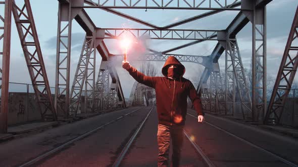 Young Man in Hoodie and Balaclava with Red Burning Signal Flare on the Road Under an Old Steel Frame