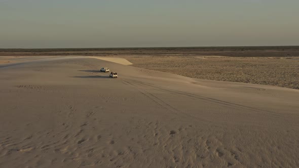 Offroad vehicles explore windy Brazil dunes with sand gusts in sunset; drone