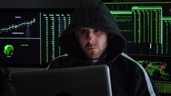 Portrait of Criminal Hooded Hacker Sitting at Desk and Breaking Into Government or Big Company Data