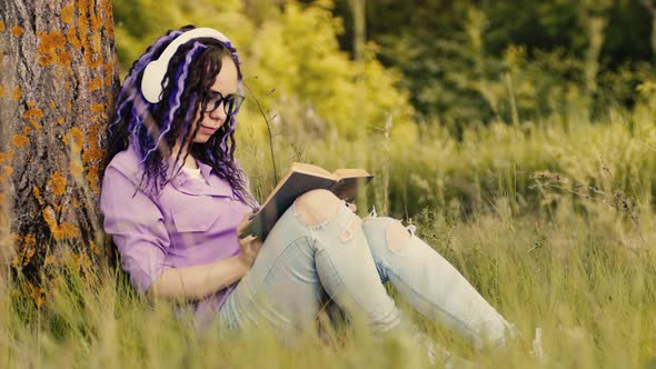 Young Woman in Glasses Headphones Reading Book Sitting Near Tree in Summertime