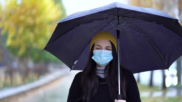 A Young Woman in a Protective Mask Walking in the Park Under Umbrella. Rainy Day, During Second Wave