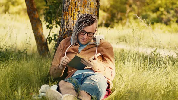 A Man in Casual Clothes and Glasses Reads Literature Under a Tree