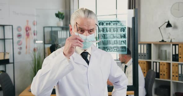 Male Doctor in Facial Mask Which Putting on His Glasses Posing on Camera in Modern Medical Office