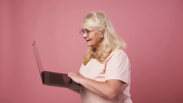 Happy Excited Senior Woman in Glasses Working on Laptop Online and Enjoying Success Pink Studio