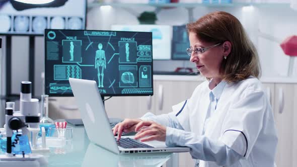 Mature Female Researcher Sits at Her Desk and Types at Laptop alt