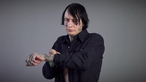 Concentrated Tattooed Young Man Adjusting Sleeves of Black Shirt Standing at Grey Background