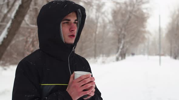 Caucasian man in black jacket with hood is warming himself in cold and drinking hot coffee.